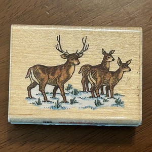 May include: Wooden stamp featuring a detailed illustration of a deer with large antlers standing with two other deer. The deer are brown with white accents, set against a snowy, grassy ground. The stamp is rectangular.