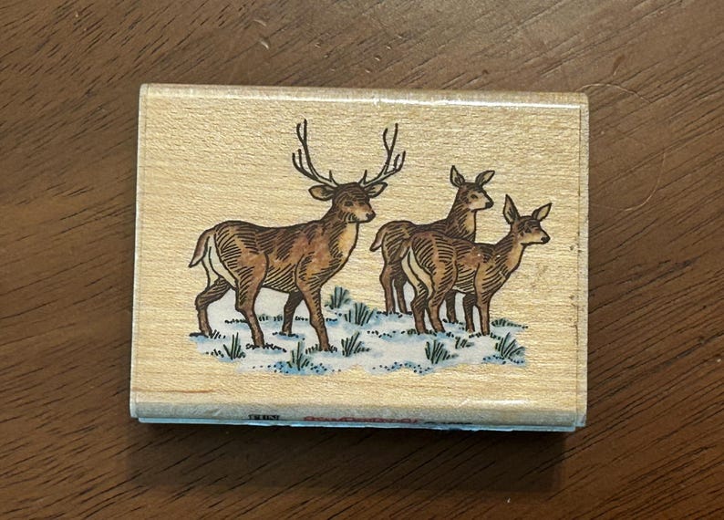 May include: Wooden stamp featuring a detailed illustration of a deer with large antlers standing with two other deer. The deer are brown with white accents, set against a snowy, grassy ground. The stamp is rectangular.