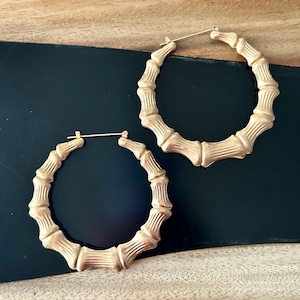 May include: A pair of gold-colored bamboo hoop earrings. The earrings have a textured surface, mimicking the look of bamboo stalks. The earrings are displayed on a black surface with a wooden background.
