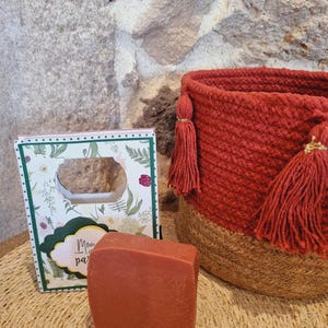 Puede incluir: Una cesta tejida roja y marrón con borlas, una tarjeta con diseños florales y el texto "Mamá, eres la mejor parte de mi día", y una barra de jabón rojo-marrón.