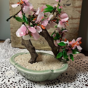 May include: A decorative bonsai tree sculpture featuring translucent pink flower petals, bright green leaves, and thin orange stamens attached to dark brown textured branches. The tree is set in a light green oval ceramic pot filled with small white stones.