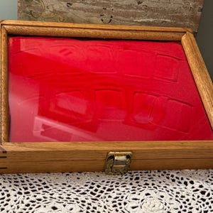 May include: A rectangular wooden display case with a clear glass lid and a vibrant red velvet interior featuring molded slots for organizing small items. The box has a brass-colored metal latch on the front and visible corner joinery details.