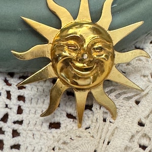 May include: A gold-colored sun-shaped brooch with a smiling face and a small, clear gem. The brooch has pointed rays and curved accents, suggesting a vintage or antique style. The sun's face has detailed features, and the overall design is ornate.