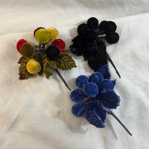 May include: Three clusters of velvet berries in various colors: yellow, red, black, and blue. Each cluster has green velvet leaves and brown stems. The berries are arranged on a white surface.