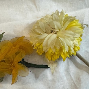 May include: Two artificial flowers on a white surface. One is a large, multi-layered flower with white and yellow petals. The other is a cluster of smaller, yellow flowers with green leaves and stems.