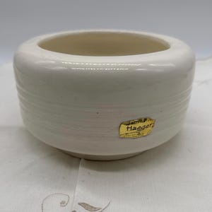 May include: A cream-colored ceramic bowl with a wide, rounded shape. The bowl has horizontal textured lines and a gold label that reads "Haeger". The interior is smooth and light-colored.