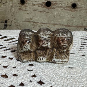 May include: A small, carved figurine depicting three monkeys sitting side-by-side. The left monkey covers its mouth, the center monkey covers its eyes, and the right monkey covers its ears. The sculpture has a weathered, textured brown and beige finish.