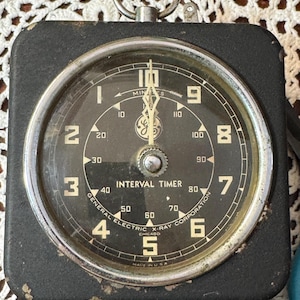 May include: A vintage black square General Electric X-Ray Corporation interval timer with a round clock face, white numerals one through ten, and a central minute scale from ten to one hundred and ten. It features a metal ring at the top.