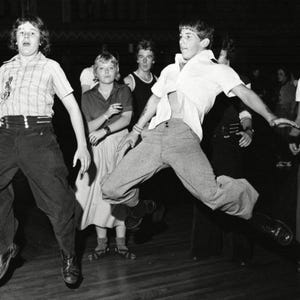 May include: A black and white photo of a group of people dancing. The people are wearing 1960s clothing and are in a dimly lit room. The people are all smiling and having fun.