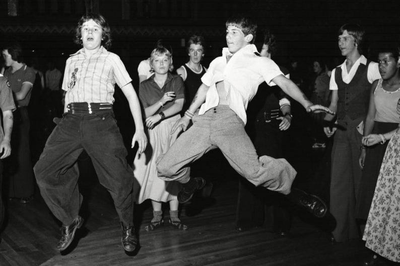 May include: A black and white photo of a group of people dancing. The people are wearing 1960s clothing and are in a dimly lit room. The people are all smiling and having fun.