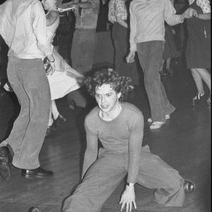 May include: A group of people dancing at a party. One person is sitting on the floor with their legs spread out in front of them. The person is wearing a sweater and pants.