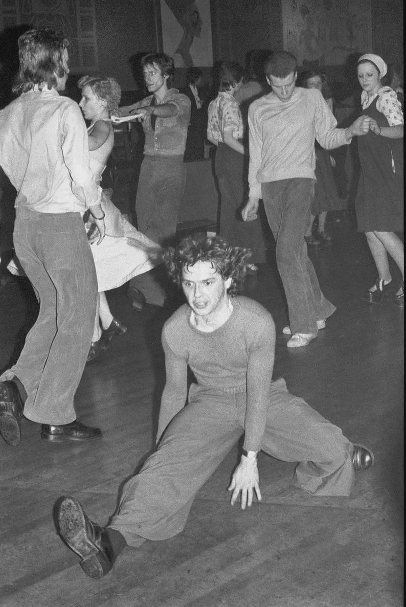 May include: A group of people dancing at a party. One person is sitting on the floor with their legs spread out in front of them. The person is wearing a sweater and pants.