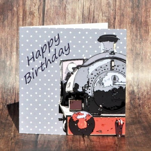 May include: A birthday card with a grey polka dot background and a steam train illustration. The card reads "Happy Birthday" in a handwritten style. The train is depicted in shades of grey, black, and red.