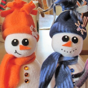 May include: Three handmade snowman decorations. Each snowman has a white body, black eyes, an orange carrot nose, and a smiling mouth. They wear colorful hats and scarves in orange, blue, and pink, with decorative buttons and snowflake accents.