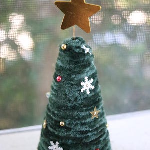 May include: A handcrafted, dark green Christmas tree decoration. The tree is cone-shaped and adorned with gold, red, and white ornaments, including stars and snowflakes. A gold star tops the tree. The tree is approximately 6 inches tall.