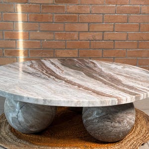 Op de afbeelding: Een ronde salontafel met een marmeren blad met bruine en witte adering. De tafel rust op twee grote, bolvormige stenen bases. Hij staat op een geweven, ronde mat, met een bakstenen muur op de achtergrond.