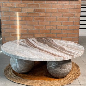 Op de afbeelding: Een ronde marmeren salontafel met een lichtbruin en wit patroon. De tafel rust op twee grote, bolvormige stenen bases. Hij staat op een geweven, cirkelvormige mat. De achtergrond omvat een bakstenen muur en een witte kast.