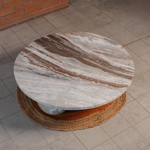 Op de afbeelding: Een ronde salontafel met een marmeren blad met bruine en witte adering. De basis is gemaakt van geweven natuurlijke vezels. De tafel heeft een diameter van ongeveer 91 cm.