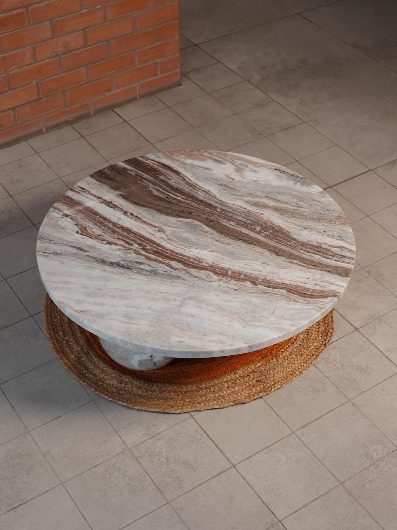 Op de afbeelding: Een ronde salontafel met een marmeren blad met bruine en witte adering. De basis is gemaakt van geweven natuurlijke vezels. De tafel heeft een diameter van ongeveer 91 cm.