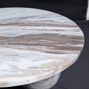 Op de afbeelding: Een ronde marmeren tafel met een wit, bruin en grijs patroon. De tafel wordt ondersteund door een afgeronde basis, wat een modern design cre&euml;ert. De natuursteen heeft een uniek aderingpatroon.