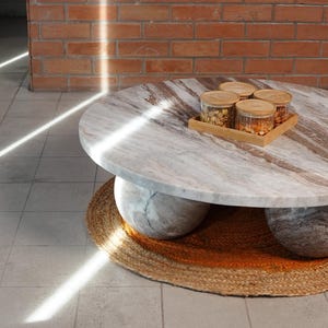 Op de afbeelding: Een ronde marmeren salontafel met een lichtbruin en grijs patroon, ondersteund door twee grote, afgeronde stenen bases. Een kleine houten dienblad met vier glazen potten staat erop. De tafel staat op een rond, geweven jute vloerkleed.