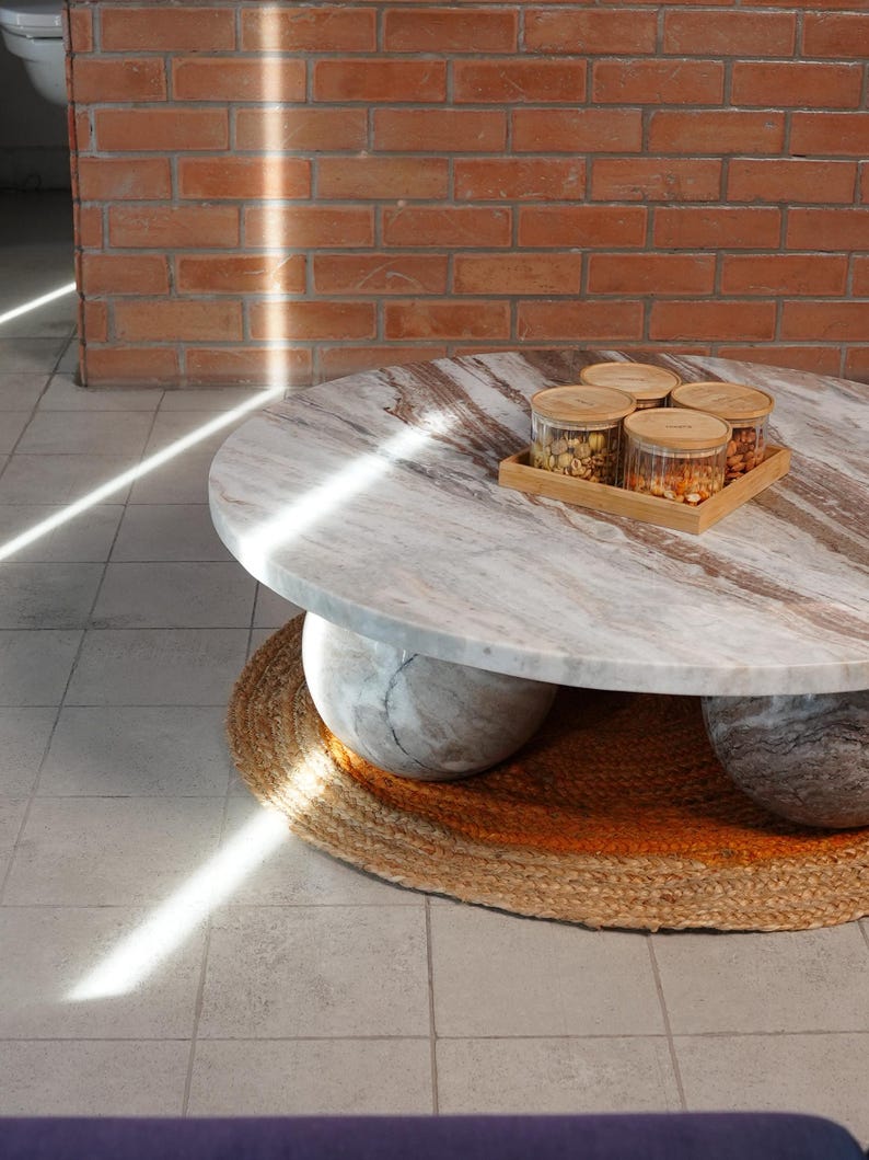 Op de afbeelding: Een ronde marmeren salontafel met een lichtbruin en grijs patroon, ondersteund door twee grote, afgeronde stenen bases. Een kleine houten dienblad met vier glazen potten staat erop. De tafel staat op een rond, geweven jute vloerkleed.
