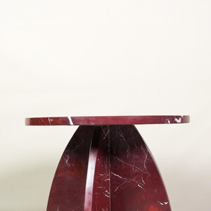 May include: A modern side table with a round top and a curved base. The table is made of red marble with white veining. The table is approximately 20 inches in diameter and 24 inches tall.