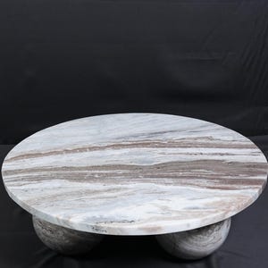Op de afbeelding: Een ronde marmeren salontafel met een wit, bruin en grijs patroon. De tafel wordt ondersteund door drie bolvormige poten. De diameter van de tafel is ongeveer 122 cm.