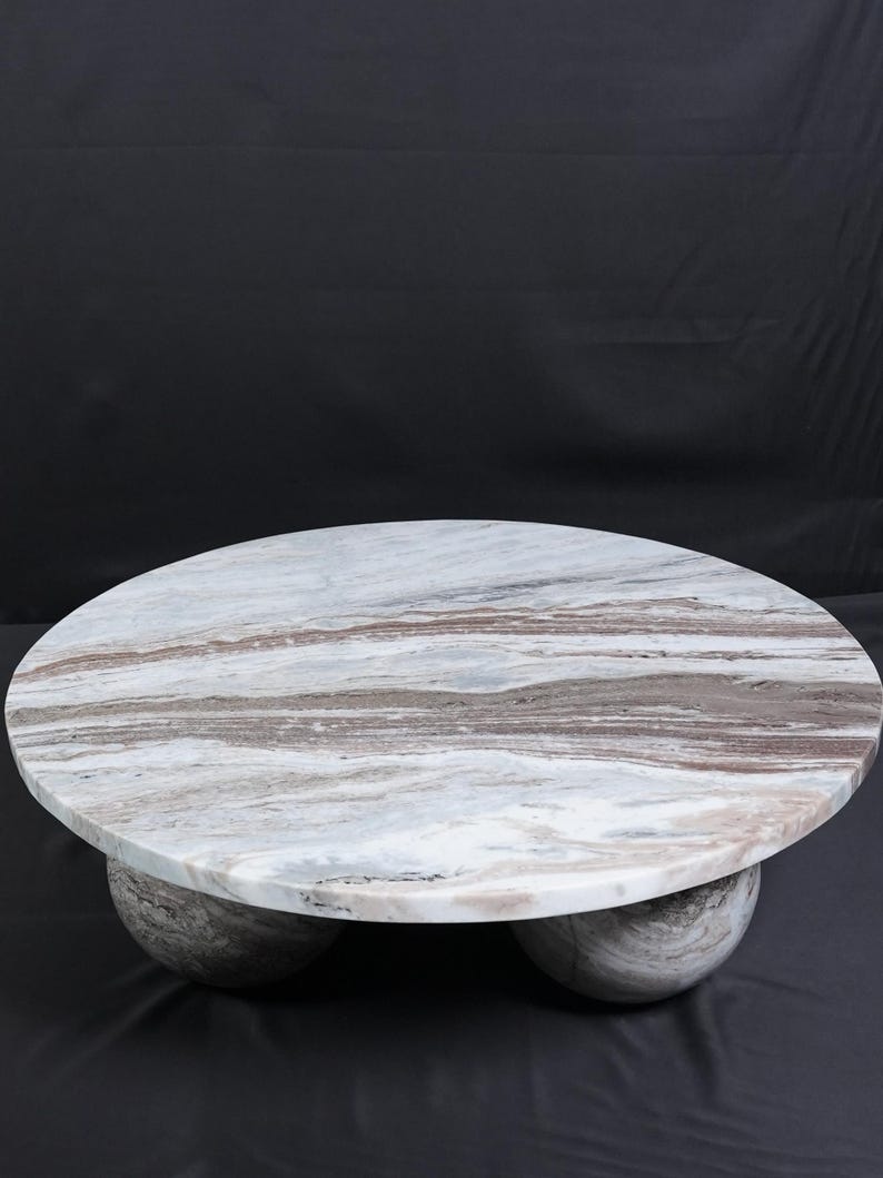 Op de afbeelding: Een ronde marmeren salontafel met een wit, bruin en grijs patroon. De tafel wordt ondersteund door drie bolvormige poten. De diameter van de tafel is ongeveer 122 cm.