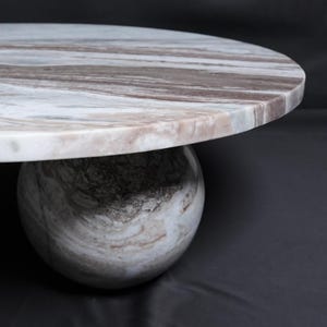 Op de afbeelding: Een ronde marmeren tafel met een blad met witte, bruine en grijze patronen. De tafel wordt ondersteund door een bolvormige voet met vergelijkbare kleurvariaties. Het ontwerp is modern en minimalistisch, geschikt voor diverse interieurstijlen.