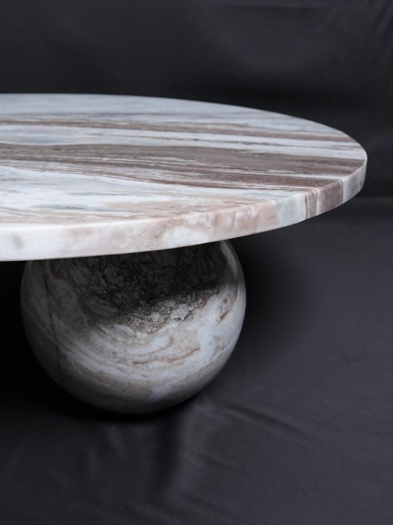 Op de afbeelding: Een ronde marmeren tafel met een blad met witte, bruine en grijze patronen. De tafel wordt ondersteund door een bolvormige voet met vergelijkbare kleurvariaties. Het ontwerp is modern en minimalistisch, geschikt voor diverse interieurstijlen.