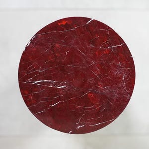 May include: A circular, red tabletop with a marbled appearance. The surface has white veining throughout, creating a natural stone effect. The table is set against a white background.