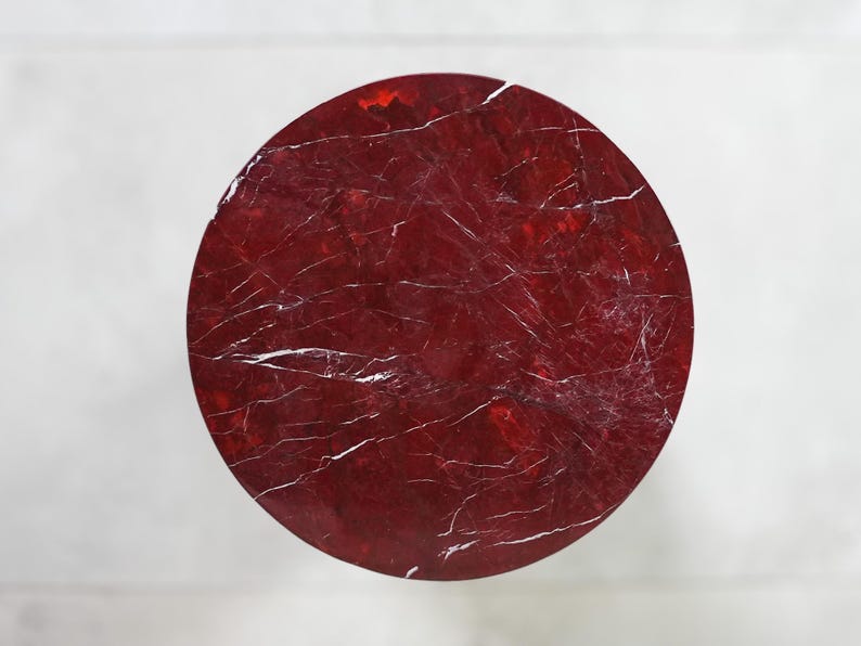 May include: A circular, red tabletop with a marbled appearance. The surface has white veining throughout, creating a natural stone effect. The table is set against a white background.