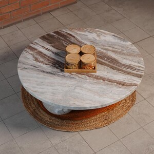 Op de afbeelding: Een ronde salontafel met een marmeren blad en een geweven basis. Het marmeren blad heeft bruine en witte adering. Vier kleine glazen potten met houten deksels staan op een houten dienblad op de tafel.