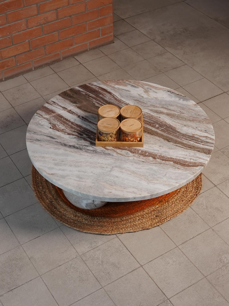 Op de afbeelding: Een ronde salontafel met een marmeren blad en een geweven basis. Het marmeren blad heeft bruine en witte adering. Vier kleine glazen potten met houten deksels staan op een houten dienblad op de tafel.