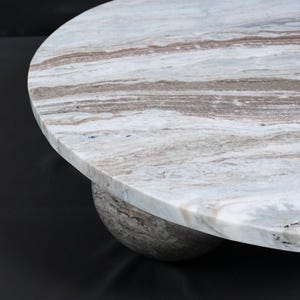 Op de afbeelding: Een ronde marmeren tafel met een lichtgrijs en bruin patroon. Het tafelblad rust op een afgeronde basis, wat zorgt voor een modern en elegant ontwerp. De tafel is geschikt voor diverse interieurstijlen.