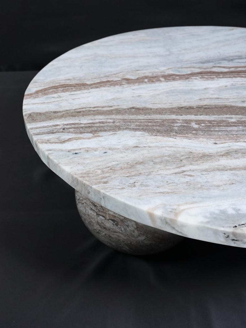 Op de afbeelding: Een ronde marmeren tafel met een lichtgrijs en bruin patroon. Het tafelblad rust op een afgeronde basis, wat zorgt voor een modern en elegant ontwerp. De tafel is geschikt voor diverse interieurstijlen.