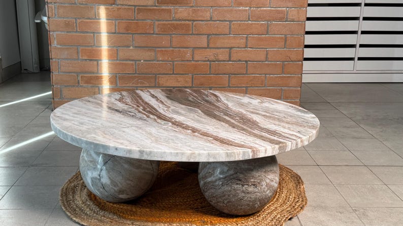 Op de afbeelding: Een ronde salontafel met een marmeren blad met bruine en witte adering. De tafel rust op twee grote, bolvormige stenen bases. Hij staat op een geweven, ronde mat, met een bakstenen muur op de achtergrond.