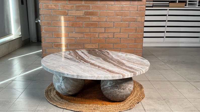 Op de afbeelding: Een ronde marmeren salontafel met een lichtbruin en wit patroon. De tafel rust op twee grote, bolvormige stenen bases. Hij staat op een geweven, cirkelvormige mat. De achtergrond omvat een bakstenen muur en een witte kast.