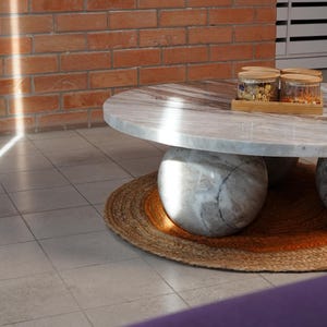 Op de afbeelding: Een ronde marmeren salontafel met een lichtgrijs en wit patroon, ondersteund door drie bolvormige stenen bases. De tafel staat op een geweven jute vloerkleed. Twee glazen potten met houten deksels staan op de tafel, bij een bakstenen muur.