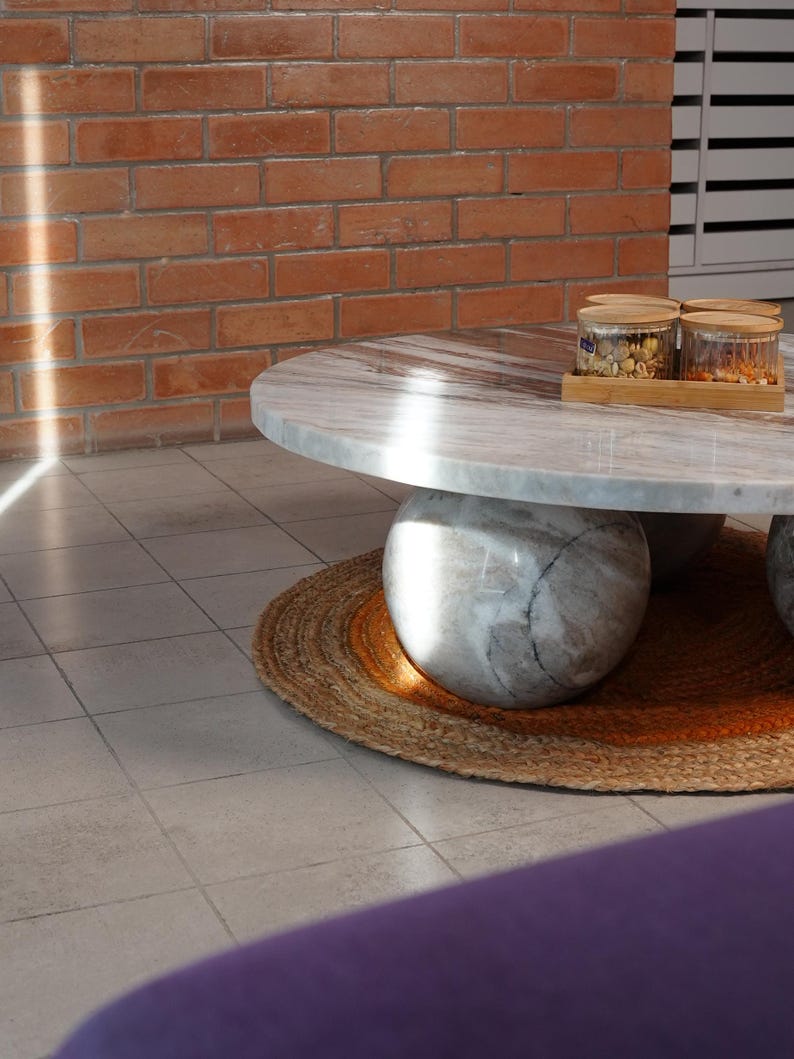 Op de afbeelding: Een ronde marmeren salontafel met een lichtgrijs en wit patroon, ondersteund door drie bolvormige stenen bases. De tafel staat op een geweven jute vloerkleed. Twee glazen potten met houten deksels staan op de tafel, bij een bakstenen muur.