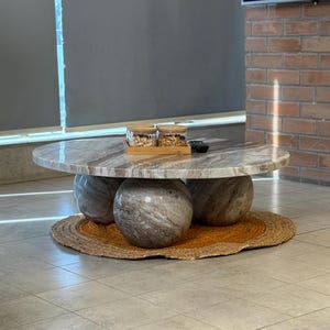Op de afbeelding: Een moderne salontafel met een ronde, marmerlook bovenkant ondersteund door drie bolvormige stenen bases. De tafel staat op een geweven jute mat. Twee glazen potten met houten deksels staan op de tafel.