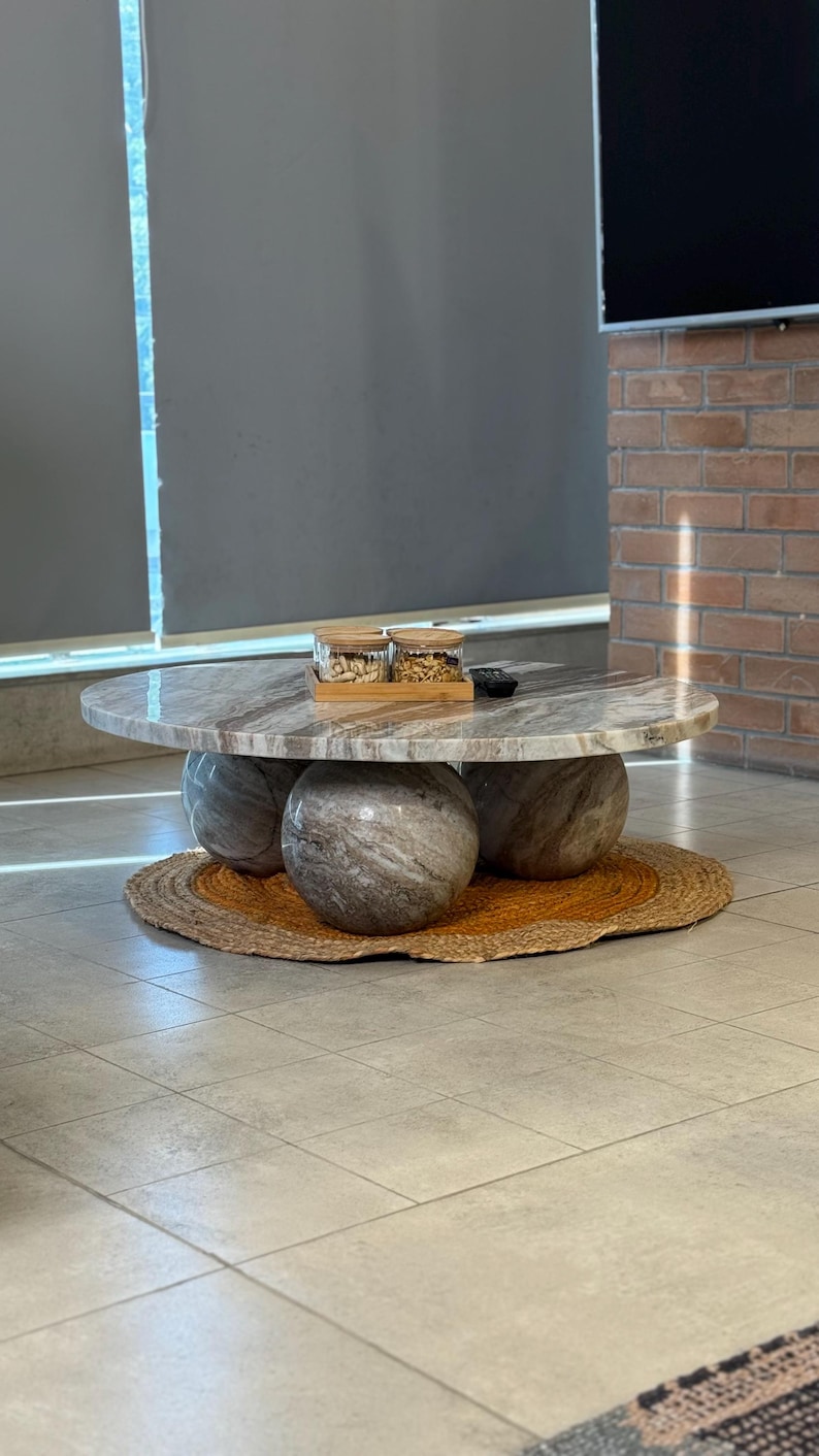 Op de afbeelding: Een moderne salontafel met een ronde, marmerlook bovenkant ondersteund door drie bolvormige stenen bases. De tafel staat op een geweven jute mat. Twee glazen potten met houten deksels staan op de tafel.