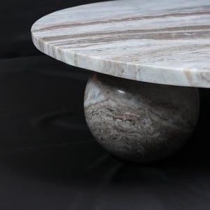 Op de afbeelding: Een ronde marmeren salontafel met een bolvormige voet. Het tafelblad is lichtgrijs met bruine aderen en de voet is van bijpassend marmer. De tafel staat tegen een donkere achtergrond.