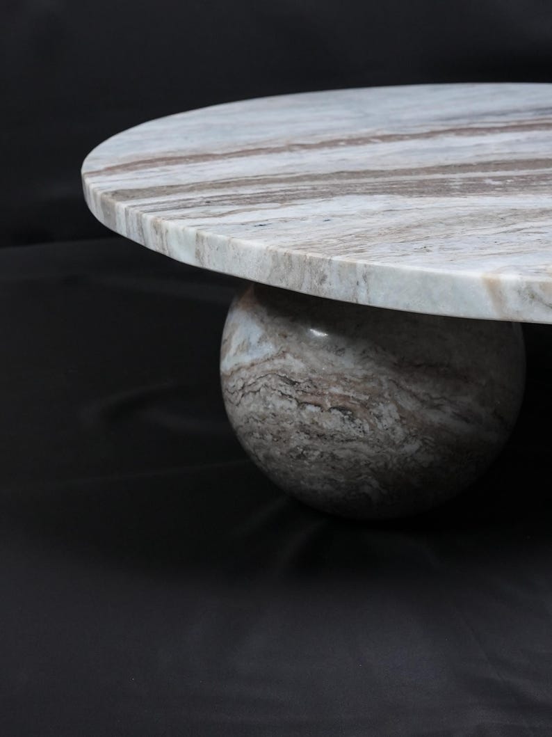 Op de afbeelding: Een ronde marmeren salontafel met een bolvormige voet. Het tafelblad is lichtgrijs met bruine aderen en de voet is van bijpassend marmer. De tafel staat tegen een donkere achtergrond.
