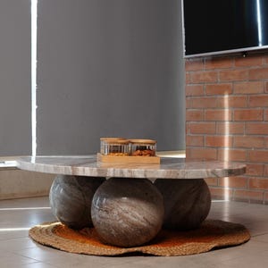 Op de afbeelding: Een moderne salontafel met een ronde marmeren bovenkant, ondersteund door drie bolvormige stenen bases. De tafel staat op een geweven jute vloerkleed met een oranje rand. Twee glazen potten met houten deksels staan erop.