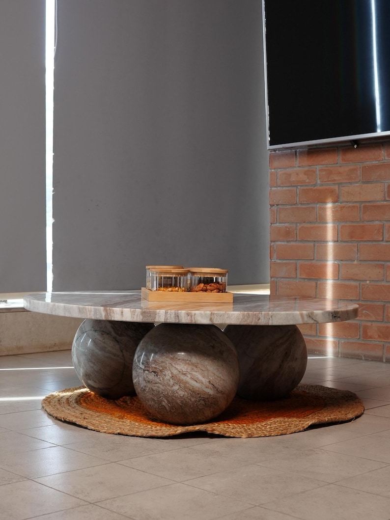 Op de afbeelding: Een moderne salontafel met een ronde marmeren bovenkant, ondersteund door drie bolvormige stenen bases. De tafel staat op een geweven jute vloerkleed met een oranje rand. Twee glazen potten met houten deksels staan erop.