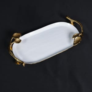 May include: A white oval serving tray with gold-colored leaf-shaped handles. The tray has a glossy finish and is set against a black background. The handles are intricately designed, adding a touch of elegance to the serving piece.