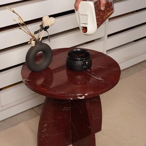 May include: A round, red marble side table with a dark gray vase, a black textured cup, and a white container with brown contents. The table has a three-legged base and a circular top. The vase holds decorative stems and a heart-shaped ornament.