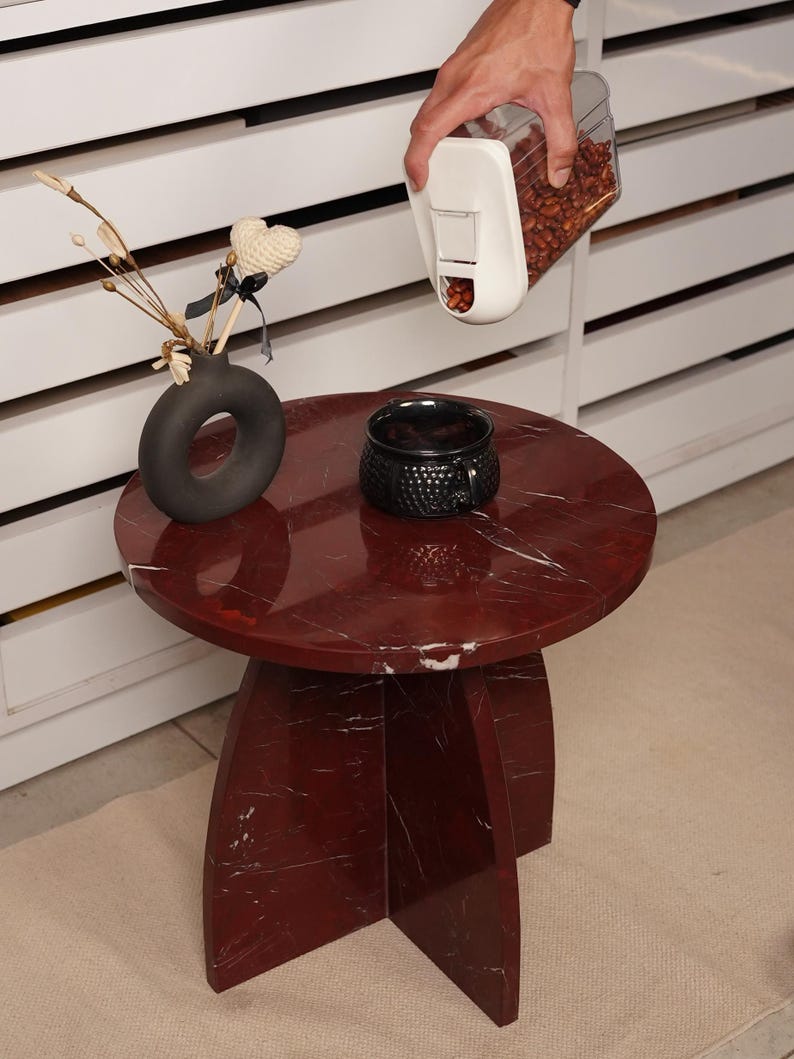 May include: A round, red marble side table with a dark gray vase, a black textured cup, and a white container with brown contents. The table has a three-legged base and a circular top. The vase holds decorative stems and a heart-shaped ornament.